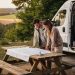 Un camping-car moderne stationné dans une aire naturelle française, avec un homme et une femme préparant leur itinéraire autour d’une table extérieure sous une douce lumière de fin d’après-midi.