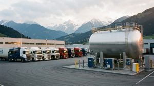 Camions poids lourds modernes stationnés dans un dépôt logistique près de Grenoble, avec une grande cuve de biocarburant et les montagnes alpines en arrière-plan.