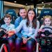 Une famille composée d’un homme, d’une femme, d’un garçon et d’une fille découvre des cockpits modernes de simulateur automobile dans un centre de loisirs intérieur.