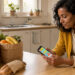 Une locataire consulte une application anti-gaspillage sur son smartphone, près de sacs de fruits et d’un pain posés sur la table lumineuse de sa cuisine.